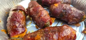 Grilled jalape&ntilde;o peppers wrapped in crispy bacon, glazed with barbecue sauce, served on a foil tray.