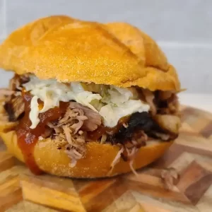 Pulled pork sandwich on a toasted bun, topped with melted cheese and barbecue sauce, served on a wooden board.