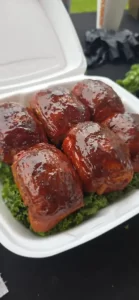 Six bacon-wrapped jalape&ntilde;o poppers glazed in barbecue sauce, served in a white takeout container on a bed of leafy greens.
