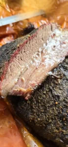 Close-up of sliced smoked brisket showing a juicy interior, smoke ring, and peppery bark, resting in a tray with rendered juices.