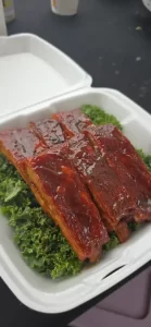 Barbecue-glazed pork ribs served in a takeout container on a bed of leafy greens.